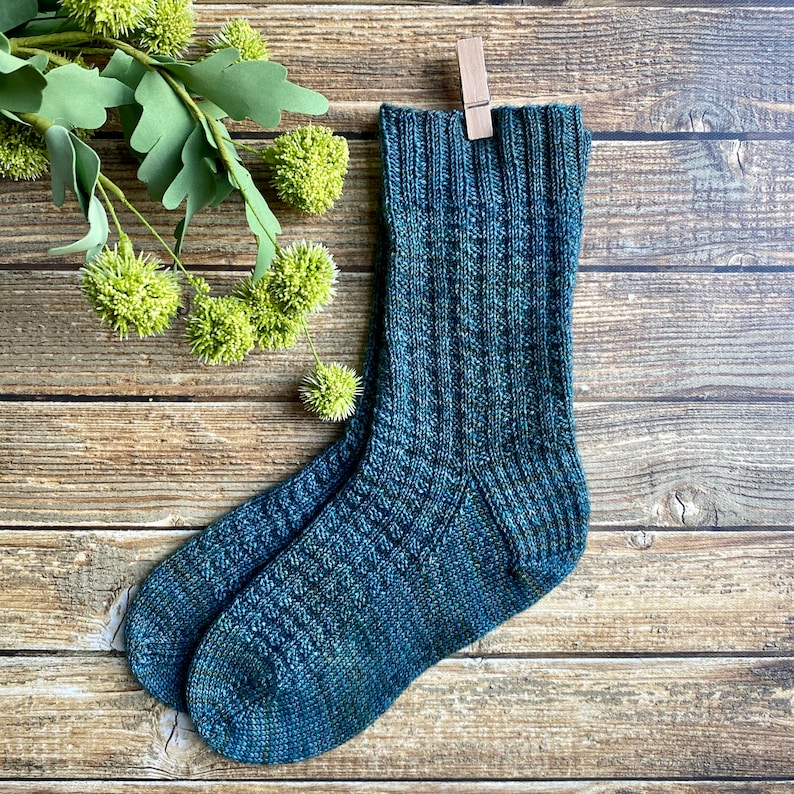 May include: A pair of blue and green knitted socks with a textured pattern. The socks are hanging on a wooden clothes peg.