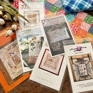 May include: A collection of cross-stitch pattern booklets featuring various designs including Sunshine On A Stem, Hello Spring, and Heart & Home, alongside a colorful patchwork fabric square and a blue plaid fabric piece with dried floral stems.