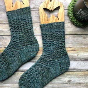 May include: Two pairs of teal blue knitted socks with a textured pattern. The socks are displayed on wooden sock blockers with butterfly cutouts.