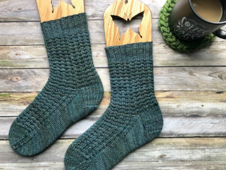 May include: Two pairs of teal blue knitted socks with a textured pattern. The socks are displayed on wooden sock blockers with butterfly cutouts.