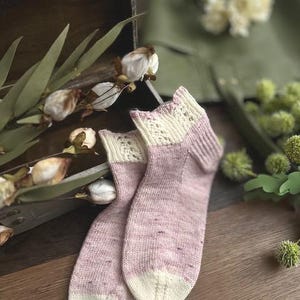 May include: A pair of pink and cream hand-knitted socks with a lacy pattern at the top. The socks are lying on a wooden surface.