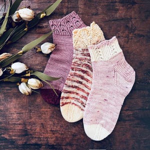 May include: Three pairs of hand-knitted socks in shades of purple, cream, and red, arranged on a dark wooden surface. The socks feature various patterns, including stripes and lace details. A sprig of dried flowers with white blooms and green leaves is placed near the socks.