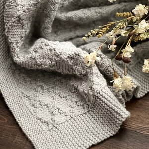 Puede incluir: Una manta de punto suave y gris con un patr&oacute;n texturizado. La manta est&aacute; doblada y drapeada sobre una superficie de madera con flores secas en la parte superior.