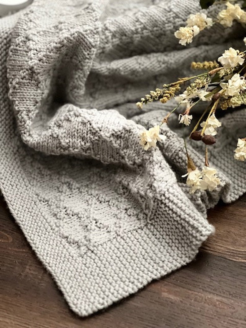 Puede incluir: Una manta de punto suave y gris con un patr&oacute;n texturizado. La manta est&aacute; doblada y drapeada sobre una superficie de madera con flores secas en la parte superior.