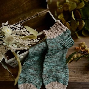 Peut inclure: Une paire de chaussettes tricotées à la main de couleur bleu sarcelle et beige avec un motif de tricot torsadé sur le bord. Les chaussettes sont posées sur une surface en bois.
