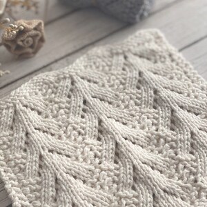 May include: A close-up of a white knitted dishcloth with a chevron pattern. The cloth is made with a soft, textured yarn.
