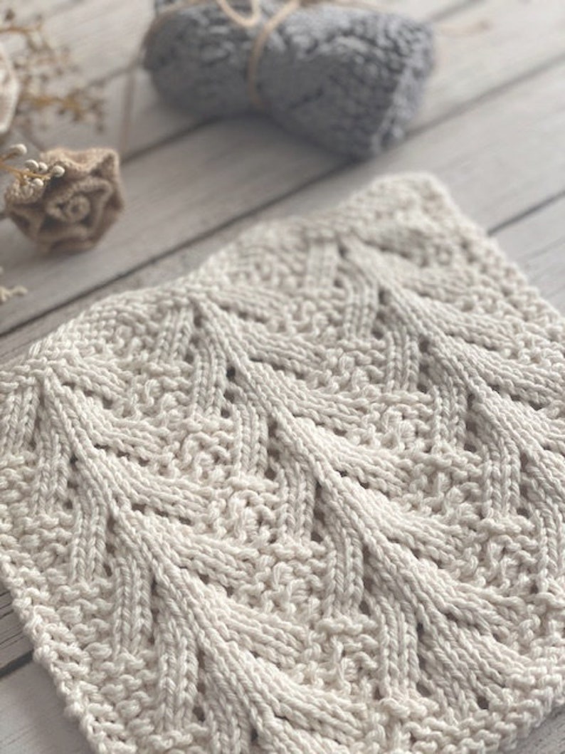 May include: A close-up of a white knitted dishcloth with a chevron pattern. The cloth is made with a soft, textured yarn.