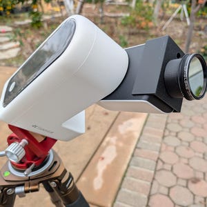 Pode incluir: Um dispositivo branco e preto com uma lente, montado em um tripé vermelho e marrom. O dispositivo tem uma tela e está inclinado para cima. A lente é preta com um anel prateado. O tripé tem pernas ajustáveis e um nível.