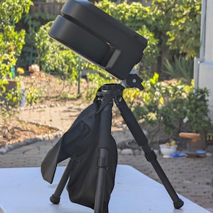 Może przedstawiać: Czarne urządzenie aparatu lub skanera zamontowane na czarnym statywie. Urządzenie ma prostokątny kształt z dwoma równoległymi sekcjami. Czarna torba transportowa jest przymocowana do statywu. Statyw stoi na białym stole, z naturalnym tłem.