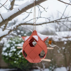 Peut inclure: Un nichoir de couleur terre cuite avec un design géométrique est suspendu à une branche d'arbre enneigée. Le nichoir présente un corps en forme de losange et des perchoirs en bois. Un petit oiseau se perche sur l'un des perchoirs en bois.
