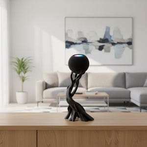 May include: A black, spherical smart speaker with a colourful light ring is displayed on a dark, sculptural stand resembling intertwined branches. The speaker is on a light wood surface in a modern living room.
