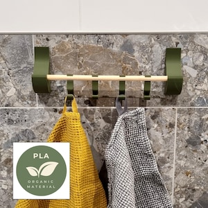 Puede incluir: Un toallero verde montado en la pared con una barra de madera natural y cuatro ganchos. Dos toallas, una amarilla y otra gris, cuelgan de los ganchos. El estante está hecho de material orgánico PLA.