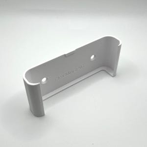 以下が含まれることがあります： 曲がった上部と2つの穴がある白いプラスチック製の壁掛け。側面には「Flex Mini 2.5G」の文字が印刷されています。マウントは、2つの垂直サポートを備えた長方形の形状をしています。