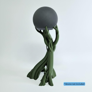 Peut inclure: Un support vert foncé en forme d'arbre avec des bras texturés en branches qui s'élèvent pour soutenir une enceinte intelligente sphérique gris foncé. La base est large et enracinée, avec une étiquette bleue *device not included.