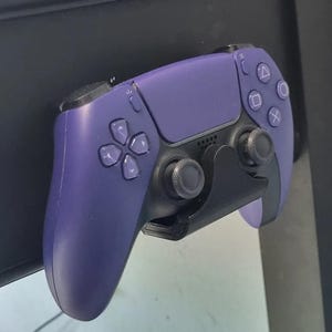 May include: A purple video game controller with black buttons and a black stand. The controller is mounted on a black surface.