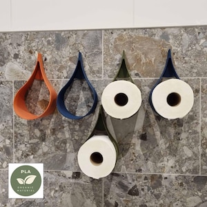 Puede incluir: Varios portarrollos de papel higiénico montados en la pared en varios colores, incluyendo naranja, azul y verde. Los soportes tienen forma de lágrima y están hechos de un material mate. Rollos de papel higiénico blanco se muestran en algunos de los soportes. La imagen también incluye un logotipo de material orgánico PLA.