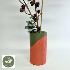 May include: A coral and olive green cylindrical vase with a geometric pattern, holding branches with pinecones and eucalyptus. The vase is made of PLA organic material. The vase is suitable for displaying flowers.