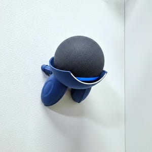 May include: A dark gray, spherical speaker sits in a navy blue holder mounted on a white wall. The holder has a curved design with three rounded supports. A thin blue accent line is visible.