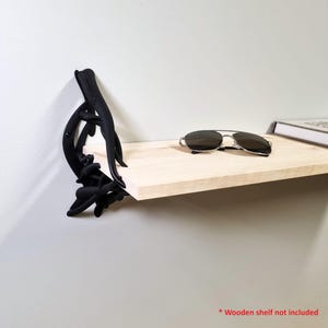 Puede incluir: Un soporte de estante de metal negro con un diseño abstracto, montado en una pared blanca. Un estante de madera de color claro está sostenido por el soporte. Gafas de sol y un libro descansan sobre el estante.
