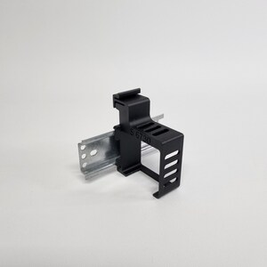 Op de afbeelding: Een zwart plastic elektrisch component met de tekst "S 6730" aan de zijkant. Het component is bevestigd aan een zilveren metalen rail met meerdere gaten. De achtergrond is effen wit.