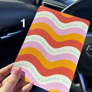 Puede incluir: Una funda de teléfono de color rosa, naranja y rojo con el texto "Spread Kindness" y "Hello Sunshine" repetido en un patrón ondulado.