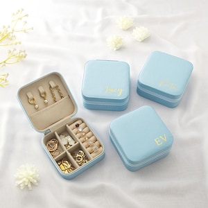 May include: A light blue jewellery box, open to reveal gold rings, earrings, and a chain necklace. Three closed light blue boxes with gold lettering are also present. The boxes are on a white surface.