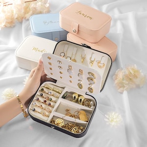 May include: An open black jewellery box displays rings, necklaces, and earrings. Several closed rectangular jewellery boxes in pastel colours are stacked behind it. The top box is peach-coloured with the name "Lauren" in gold script. The jewellery is gold-toned.
