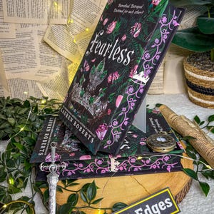 May include: A stack of books with the title "Fearless" by Lauren Roberts, featuring a dark cover with floral and crown designs. A decorative sword, a pocket watch, and a sign that says "Storybound Edges" are also present.