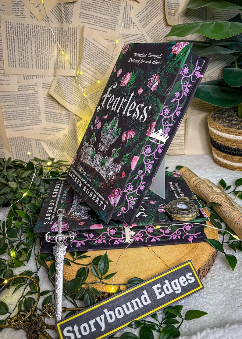 May include: A stack of books with the title "Fearless" by Lauren Roberts, featuring a dark cover with floral and crown designs. A decorative sword, a pocket watch, and a sign that says "Storybound Edges" are also present.