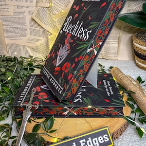 May include: Stacked copies of the book "Reckless" by Lauren Roberts, featuring a black cover with red floral and leaf accents. The title is in white script. A decorative sword and a sign that says "Storybound Edges" are also visible.