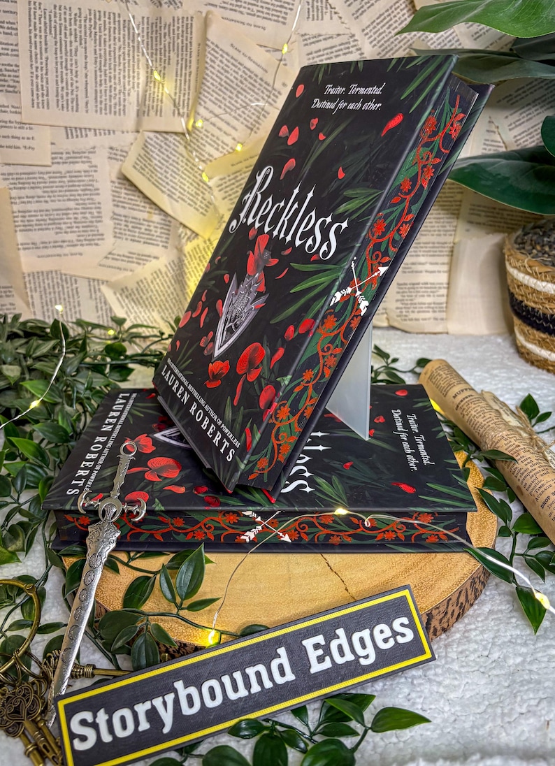 May include: Stacked copies of the book "Reckless" by Lauren Roberts, featuring a black cover with red floral and leaf accents. The title is in white script. A decorative sword and a sign that says "Storybound Edges" are also visible.