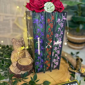 May include: A decorative book stack with red roses and a green flower on top. The book spines are black with colorful floral and sword designs. A small bear statue and a sign that says "Storybound Edges" are in the foreground.