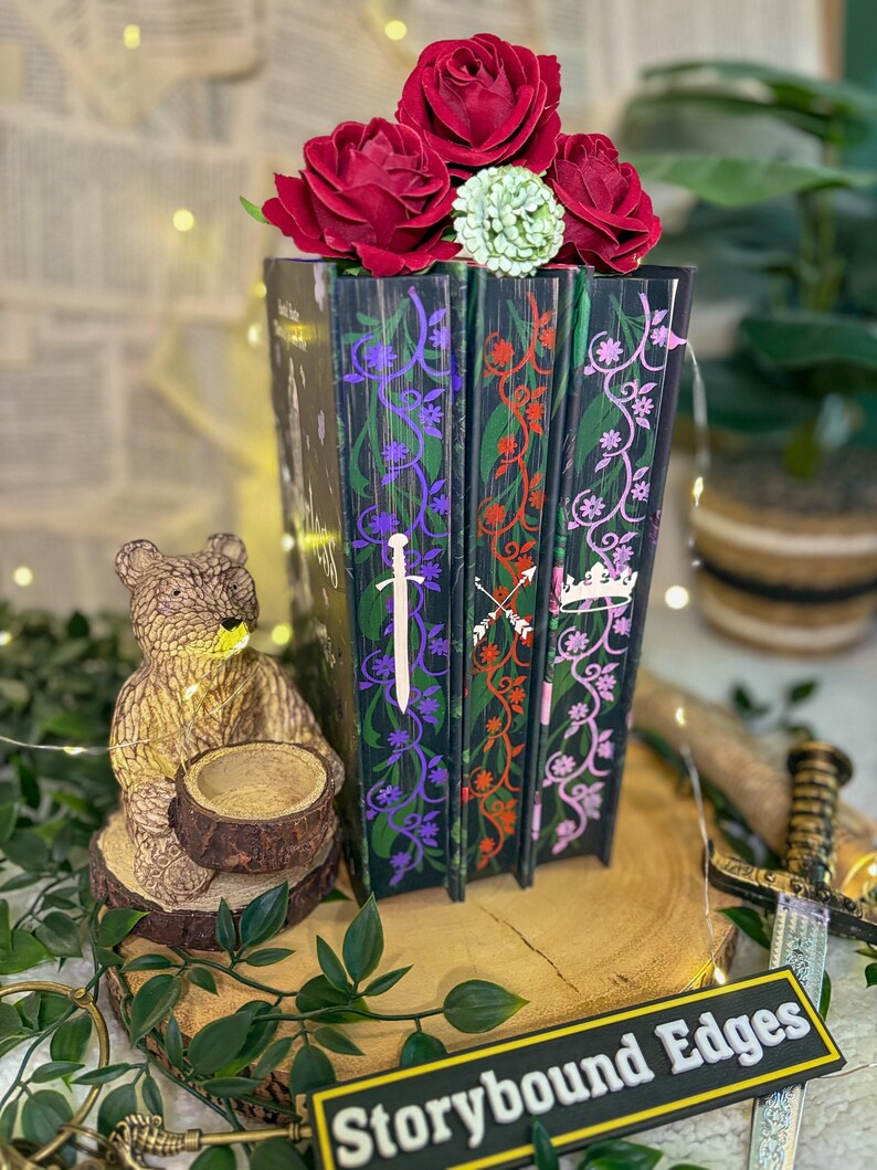 May include: A decorative book stack with red roses and a green flower on top. The book spines are black with colorful floral and sword designs. A small bear statue and a sign that says "Storybound Edges" are in the foreground.