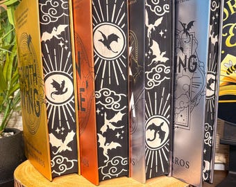 May include: A collection of five hardcover books with metallic gold, copper, and silver spines. The page edges feature black and white printed designs of dragons, clouds, and sunbursts. The text Fourth Wing and Iron Flame is visible on the spines.