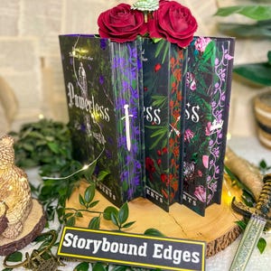 May include: A display of four hardcover books with floral and fantasy-themed covers. The books are arranged on a wooden surface, adorned with greenery and red roses. The title "Powerless" is visible on one book. A sign reads "Storybound Edges".