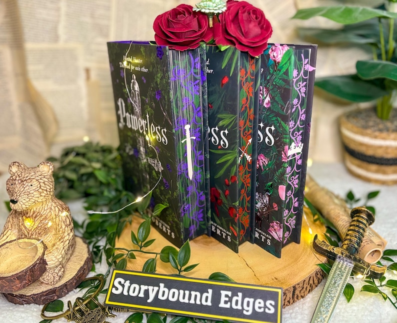 May include: A display of four hardcover books with floral and fantasy-themed covers. The books are arranged on a wooden surface, adorned with greenery and red roses. The title "Powerless" is visible on one book. A sign reads "Storybound Edges".