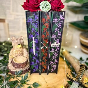 May include: A decorative set of three black books with floral and sword designs, topped with red roses and a green accent. The base features the text "Storybound Edges".
