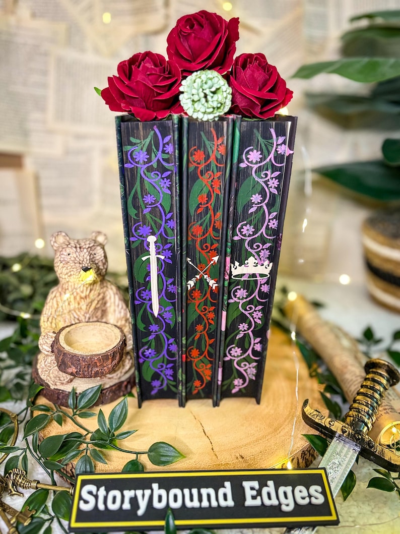 May include: A decorative set of three black books with floral and sword designs, topped with red roses and a green accent. The base features the text "Storybound Edges".