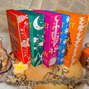 Könnte beinhalten: Ein Set aus sechs Fantasy-Romanen mit bunten Covern, die Illustrationen von Blumen, einer Mondsichel, einem Schwert, Schneeflocken und Flammen zeigen. Die Bücher tragen die Titel "A Court of Thorns and Roses" und "A Court of Mist and Fury".