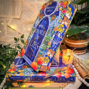 Könnte beinhalten: Zwei gebundene Bücher mit dem Titel "A Dance of Lies" von Brittney Arena. Die Bücher haben einen blauen Einband mit floralen und Buntglas-Designs. Das Bild enthält eine brennende Kerze, dekorative Schere und ein Schild mit der Aufschrift "Storybound Edges".