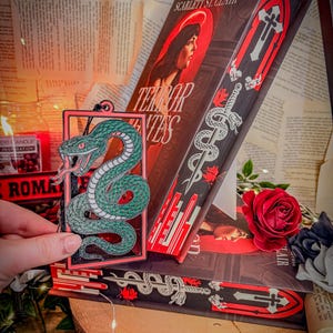 Op de afbeelding: Een boek getiteld "Terror Tentes" van Scarlett St. Clair, met een rood-zwart omslagontwerp. Een groen slangvormig bladwijzer wordt voor het boek gehouden. Een rode roos en lichtslingers zijn ook zichtbaar.