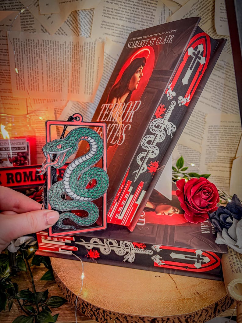 Op de afbeelding: Een boek getiteld "Terror Tentes" van Scarlett St. Clair, met een rood-zwart omslagontwerp. Een groen slangvormig bladwijzer wordt voor het boek gehouden. Een rode roos en lichtslingers zijn ook zichtbaar.