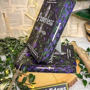May include: Stacked copies of the book "Powerless" by Lauren Roberts. The book covers are black with purple and green floral designs and a white sword. The title is in white script. The book is displayed on a wooden surface with greenery and fairy lights.