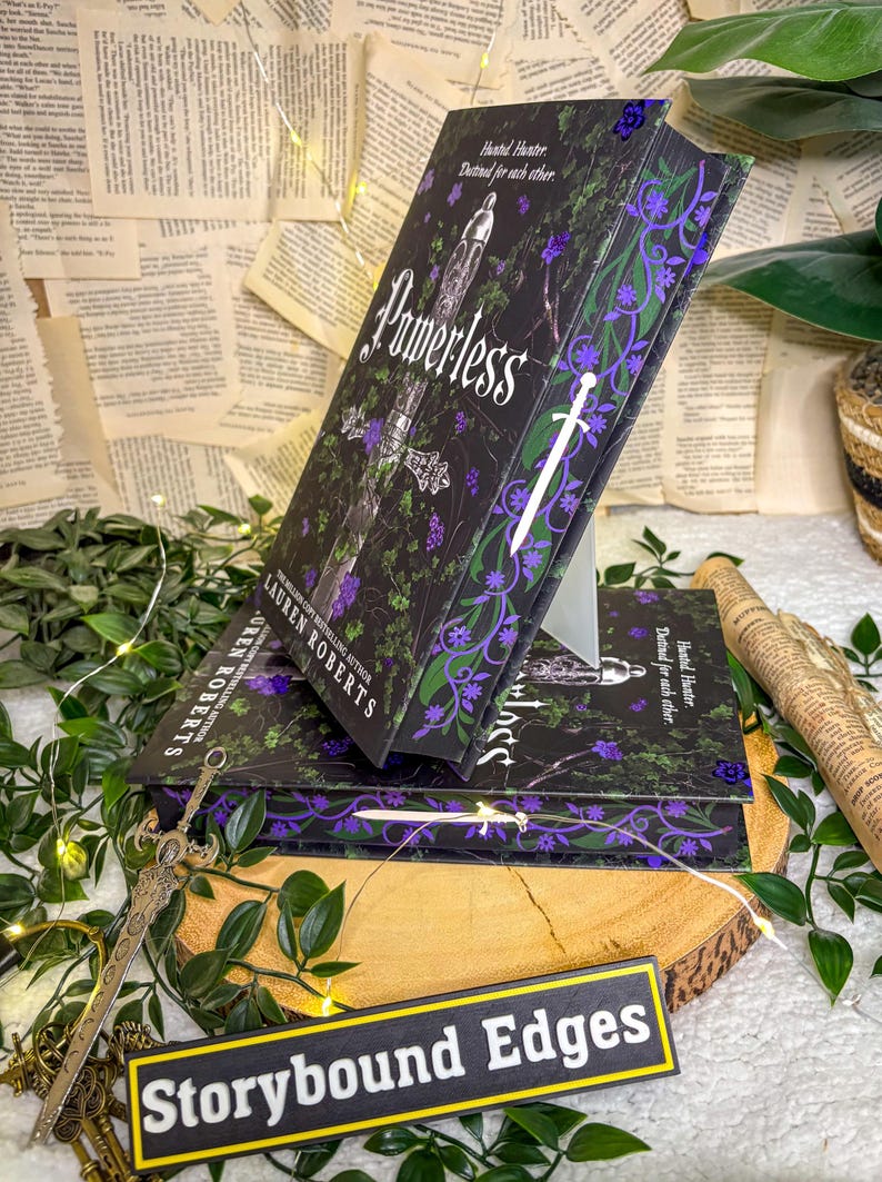 May include: Stacked copies of the book "Powerless" by Lauren Roberts. The book covers are black with purple and green floral designs and a white sword. The title is in white script. The book is displayed on a wooden surface with greenery and fairy lights.