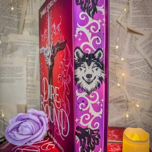 Op de afbeelding: Een hardcover boek getiteld "Dire Round" van Sable Sorensen. De omslag is een verloop van rood en roze met een zilveren illustratie van een zwaard. De rug is paars met een wolf illustratie en witte krullen. Een paarse roos en een kaars staan op de voorgrond.