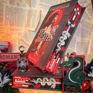 Op de afbeelding: Een stapel boeken, waaronder "Terror at the Gates" van Scarlett St. Clair, met een portret van een vrouw op de cover. Een decoratieve bladwijzer met een slangenontwerp is zichtbaar. Een decoratieve parfumfles en een groene slangvormige bladwijzer zijn ook aanwezig.
