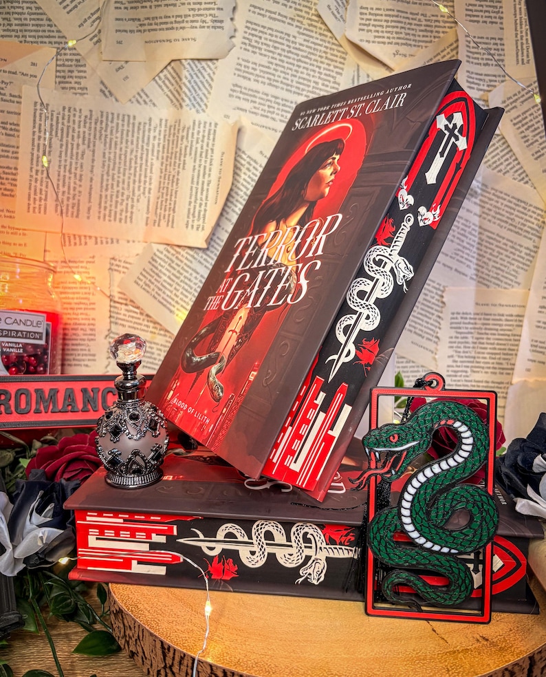 Op de afbeelding: Een stapel boeken, waaronder "Terror at the Gates" van Scarlett St. Clair, met een portret van een vrouw op de cover. Een decoratieve bladwijzer met een slangenontwerp is zichtbaar. Een decoratieve parfumfles en een groene slangvormige bladwijzer zijn ook aanwezig.