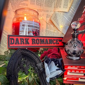 Op de afbeelding: Een brandende Yankee Candle-pot met een "Cherry Vanilla" label staat naast een rood bord met de tekst "DARK ROMANCE". Een decoratieve parfumfles en een gotische boog zijn ook zichtbaar, samen met rode en witte rozen en een boek.