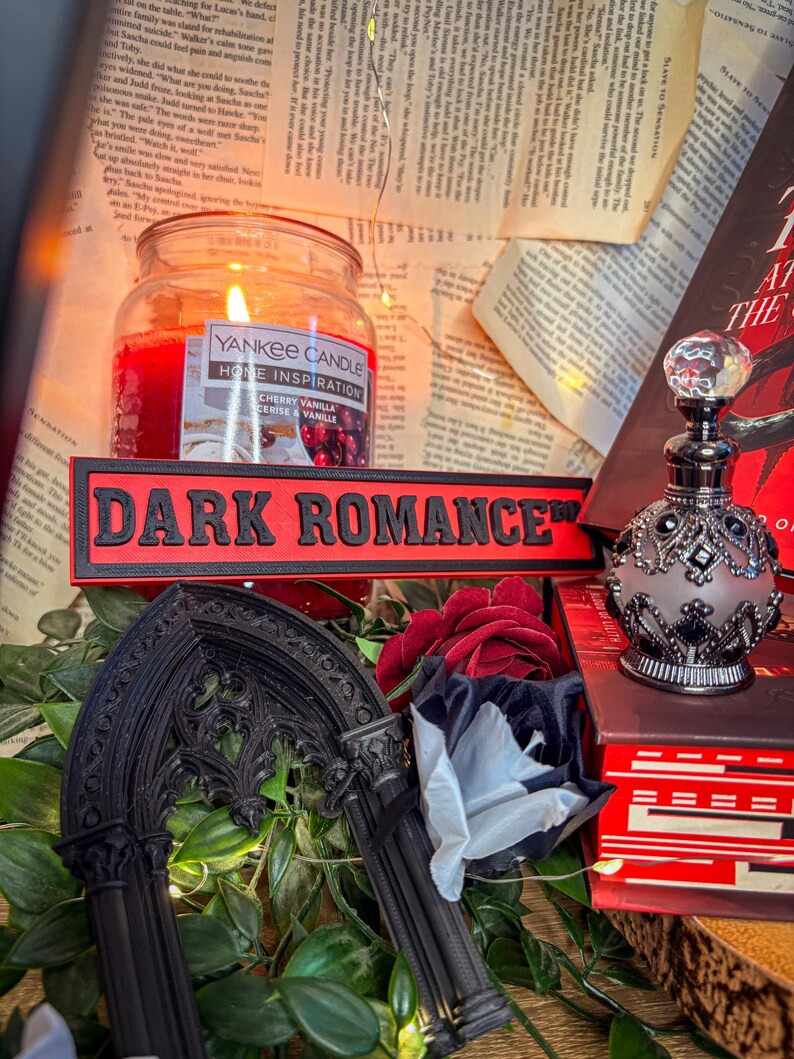 Op de afbeelding: Een brandende Yankee Candle-pot met een "Cherry Vanilla" label staat naast een rood bord met de tekst "DARK ROMANCE". Een decoratieve parfumfles en een gotische boog zijn ook zichtbaar, samen met rode en witte rozen en een boek.