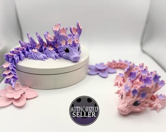 May include: Two purple and pink dragon figurines with flower details. The dragons are made of a soft, pliable material and have a whimsical, fairytale-like appearance.
